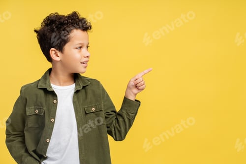 Preview: Smiling African American boy standing, pointing to his side in studio, isolated on yellow background