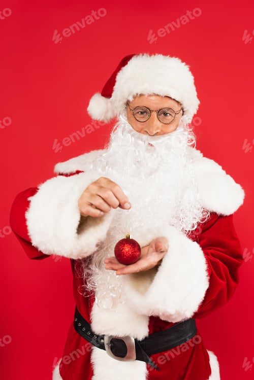 Preview: Santa claus in costume and eyeglasses holding christmas ball isolated on red
