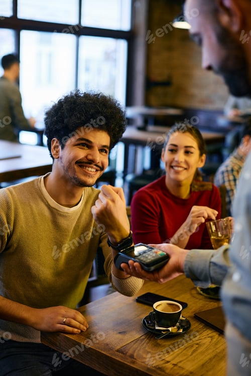 Preview: Happy man using smart watch while making payment to a waiter in a pub.