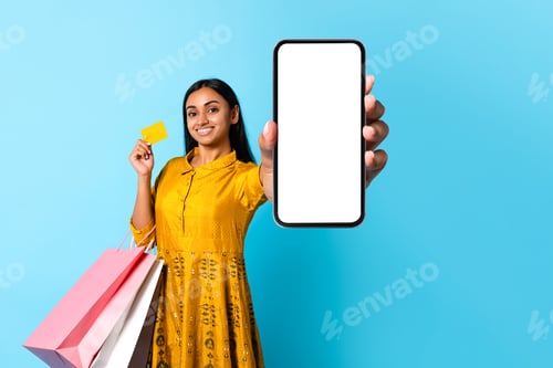 Preview: indian woman shopaholic holds cellphone displaying empty screen, blue background
