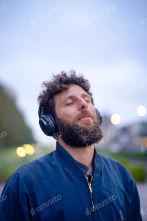 Preview: Bearded man enjoying music with closed eyes wearing black headphones outdoors at dusk.