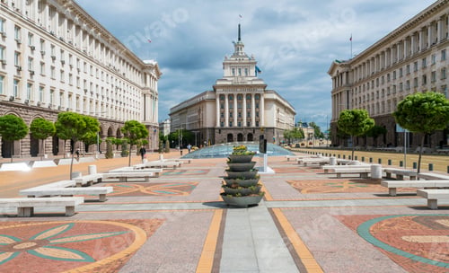 Preview: Sunny day at a grand plaza with ornate pavement and a fountain leading to a majestic building.