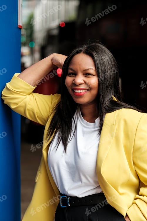 Preview: Smiling black businesswoman leaning against blue pillar in urban setting