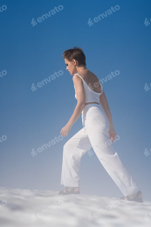 Preview: Slim blond woman walks through snow with light fog. Female in white crop top and pants, sandals