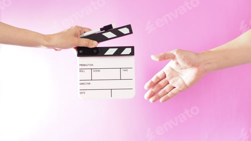 Preview: Hand is holding clapper board and marker pen and sending to ather hand on pink background.