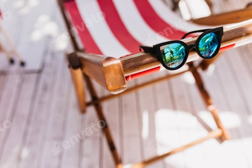 Preview: Cute female glasses lying on deck chair. Photo of black summer sunglasses on striped recliner.