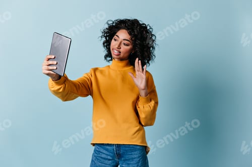 Preview: Appealing girl waving hand during video call. Blissful curly woman using digital tablet.