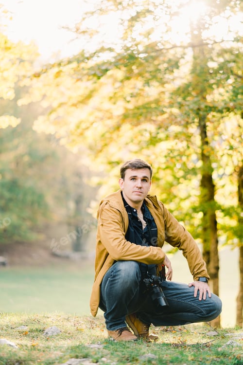 Preview: Young man with a camera around his neck squatted down in the autumn forest