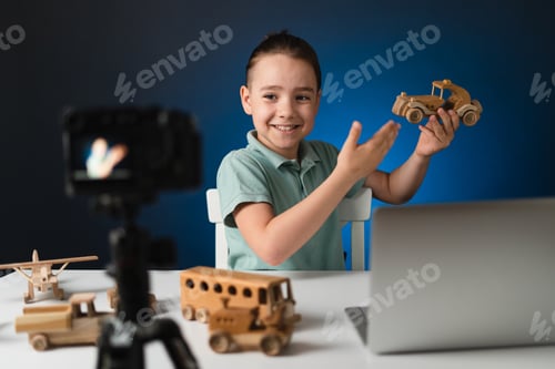 Preview: Kid blogger filming video on camera using the laptop, indoors. Promoting eco wooden toys.