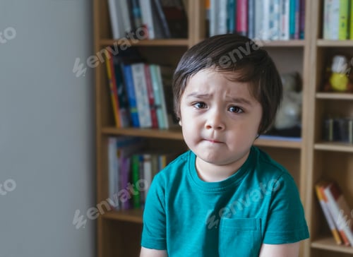 Preview: sad kid looking at camera with unhappy face, Angery little boy sitting alone with blurry bookshelf