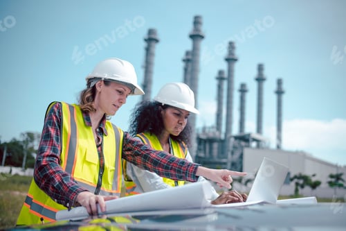 Preview: Engineers wearing safety gear, including hard hats examining survey a large blueprint tablet standin