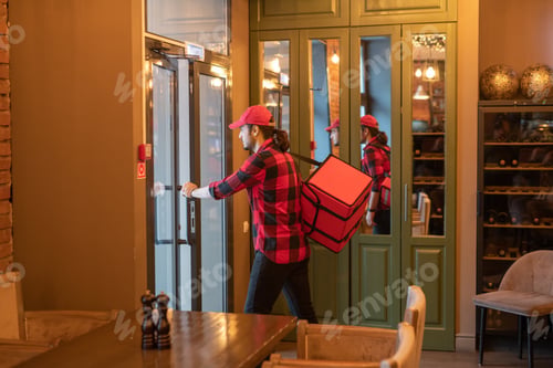 Preview: Young busy courier with big bag opening the door while going to deliver orders