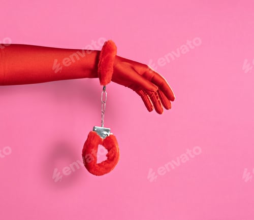 Preview: Female hand in red gloves with red fluffy handcuffs on pink background.