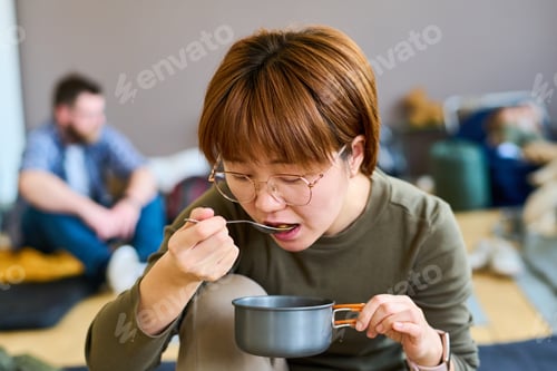 Preview: Young hungry Asian female refugee with small pan eating hot food
