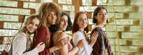 Preview: joyful, stylish multiethnic girlfriends smiling at camera and embracing in retreat center, banner