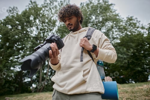 Preview: positive indian man holding digital camera and standing with backpack in forest, photographer
