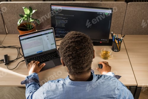 Preview: Black Man Writing Lines of Code On Desktop PC With Two Monitors and a Laptop Aside in Stylish Office