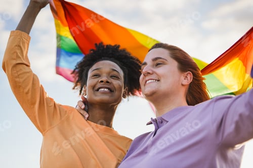 Preview: Young lesbian multiracial couple holding LGBT rainbow flag outdoor at pride parade