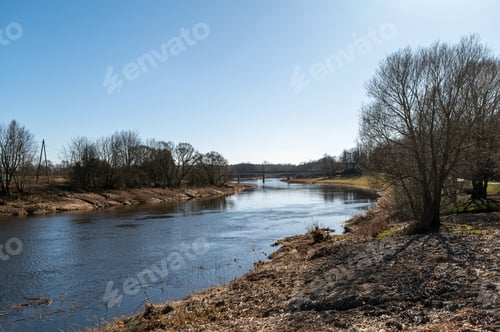 Preview: Riverbank under clear blue sky with distant bridge and trees along the shoreline
