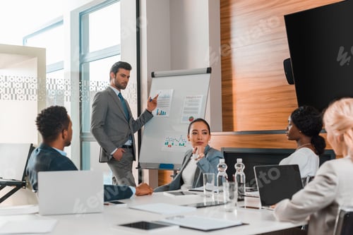 Preview: handsome businessman pointing with finger at flipchart with infographics while multicultural