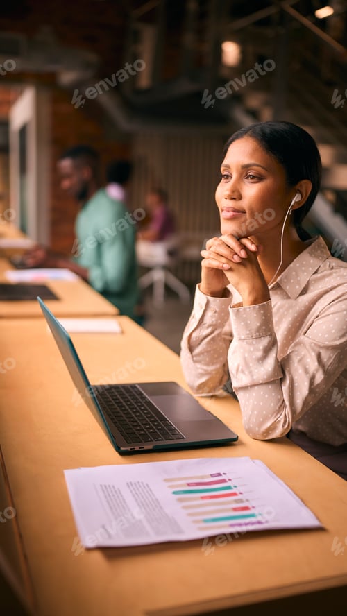 Preview: Businesswoman Working In Modern Multi-Cultural Office Listening To Music On Earphones Or Earbuds