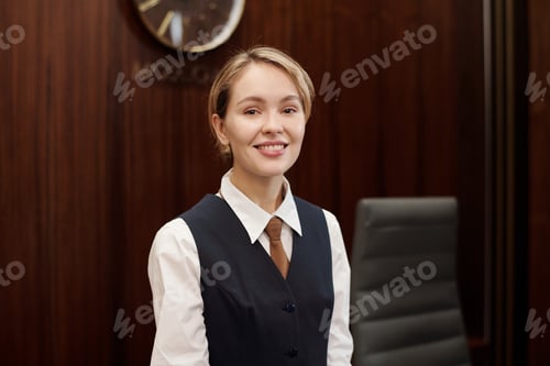 Preview: Happy young blond receptionist in uniform looking at camera by workplace