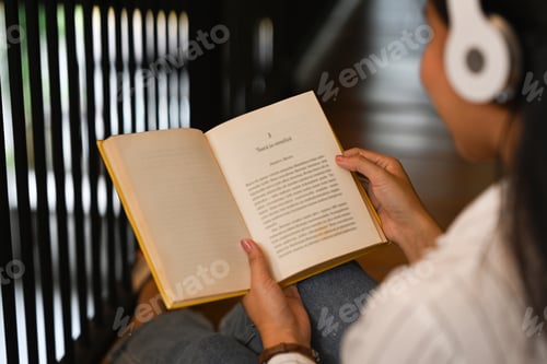 Preview: Over shoulder view of young woman wearing headphone and reading book in library.
