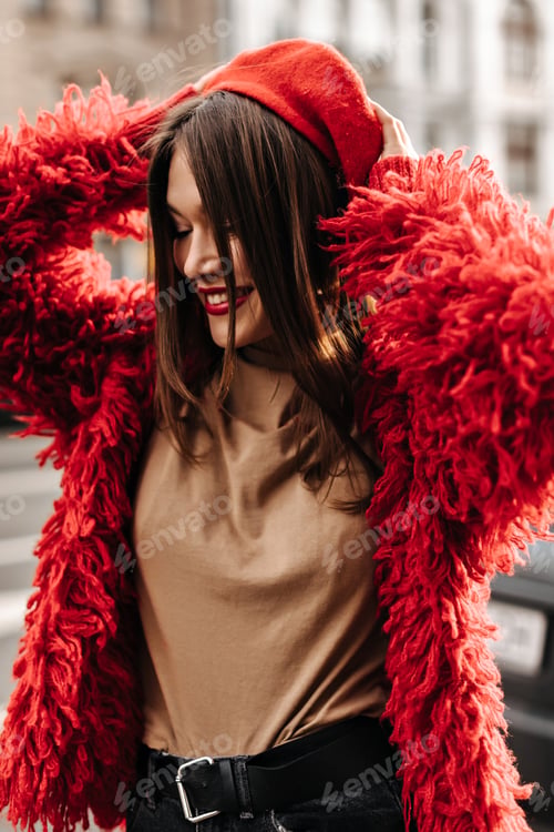 Preview: Fashionable girl in beige top and red coat puts on beret in French style, walking around city