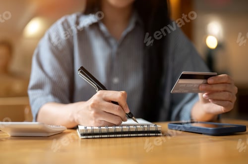 Preview: Close up a woman holding credit or debit card and writing with pen in notebook sitting at cafe table