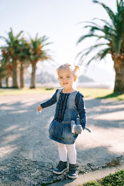 Preview: Little smiling girl stands on the road near palm trees with a toy in her hand
