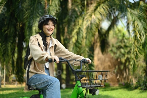 Preview: Smiling young woman in safety helmet riding a bicycle through city spring park
