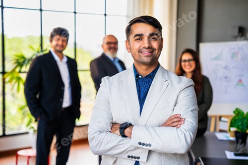 Preview: Confident Indian business leader standing with folded hands, colleagues out of focus in the office