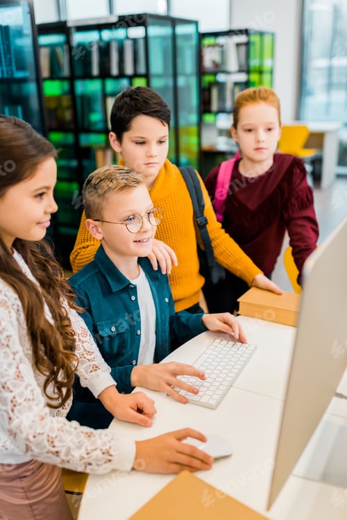 Preview: high angle view of adorable smiling schoolkids using desktop computer in library