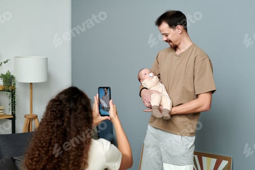 Preview: Happy young man looking at baby on his hands while female taking photo of them