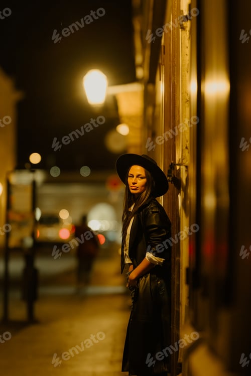 Preview: Stylish woman wearing black hat leaning against wall at night in city street