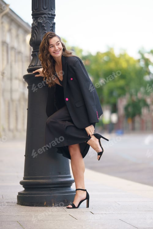 Preview: Woman in a black blazer and heels, playfully leaning against a lamp post on a city street, smiling w