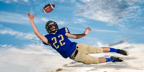 Preview: Male teenage American football player reaching to catch ball mid air against blue sky