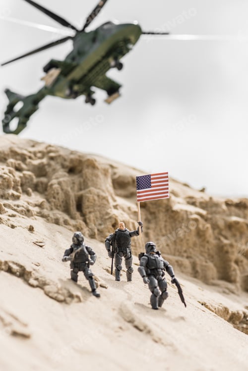 Preview: selective focus of toy soldiers on sand hill with american flag and helicopter on background