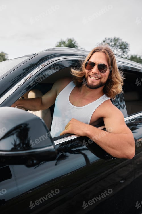Preview: Young man in sunglasses smiling from a black car window on a sunny day