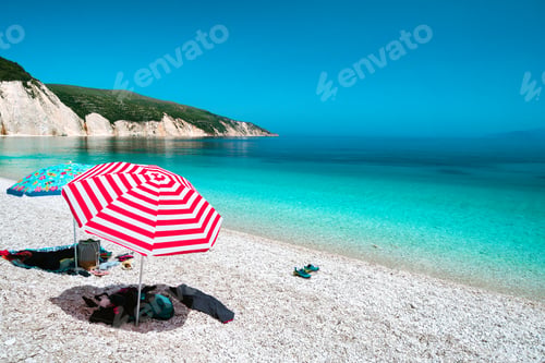 Preview: Sun beach umbrellas on a pebble beach with azure blue calm sea, white rocks and clear sky in