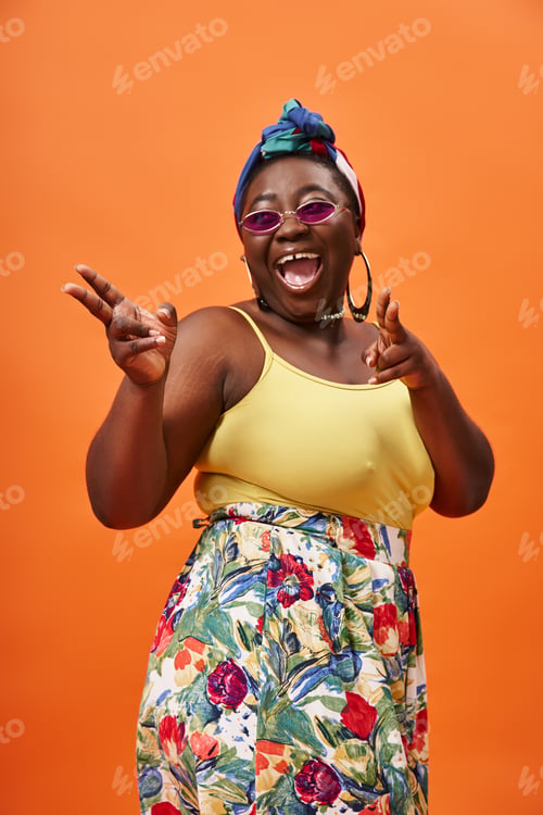 Preview: excited african american plus size woman with headscarf and sunglasses posing on orange backdrop