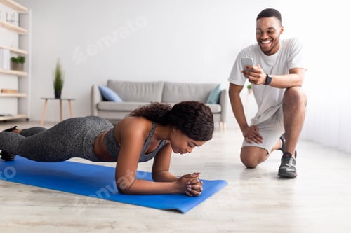 Preview: Young black couple working out as team at home, fit lady standing in elbow plank, boyfriend noting
