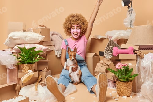 Preview: Positive joyful woman with curly hair applies beauty mask poses with pet on floor around cardboard b