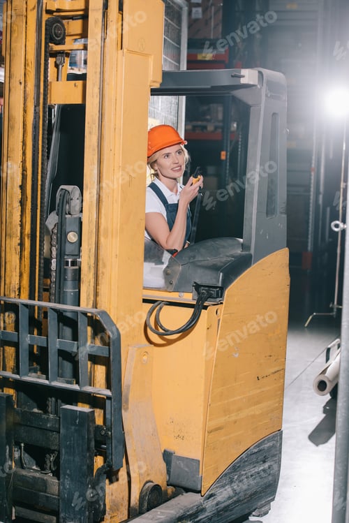 Preview: smiling workwoman sitting in forklift loader, talking on walkie talkie and looking away