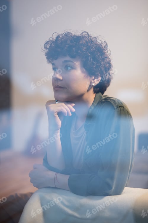 Preview: Young relaxed brunette female in casualwear sitting on couch by window