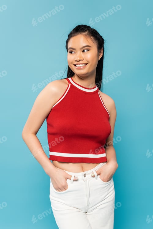 Preview: joyful asian woman in red top looking away while holding hands in pockets of white pants isolated on