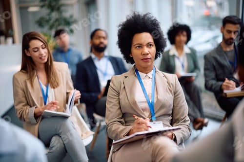 Preview: Black female manager writing in notebook during business seminar in conference hall.