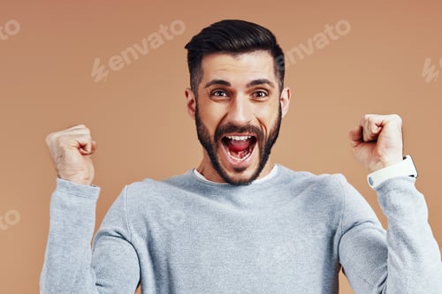 Preview: Happy young man in casual wear cheering and smiling while standing against brown background