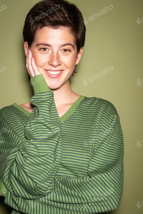 Preview: portrait of joyful brunette woman in striped pullover touching face and looking at camera on green