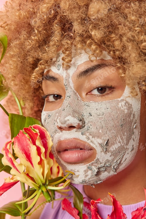 Preview: Vertical shot of serious woman with curly hair applies natural facial mask for skin peeling looks di